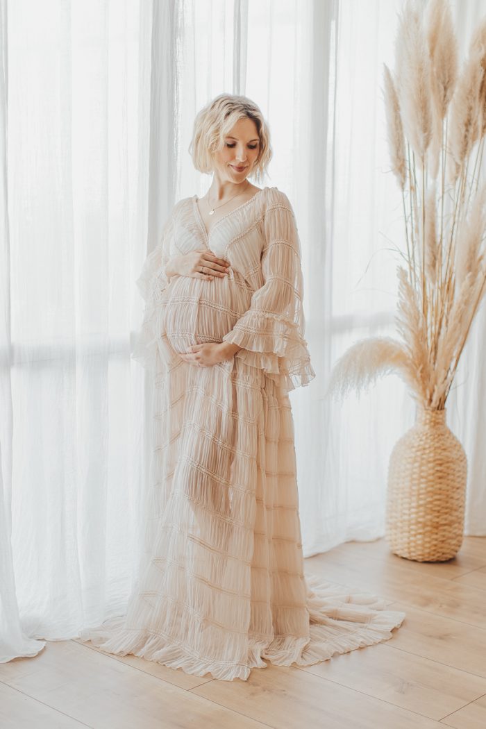 Schwangere Frau in einem boho-romantischen Kleid im Tageslicht-Studio bei einem Babybauch-Fotoshooting.